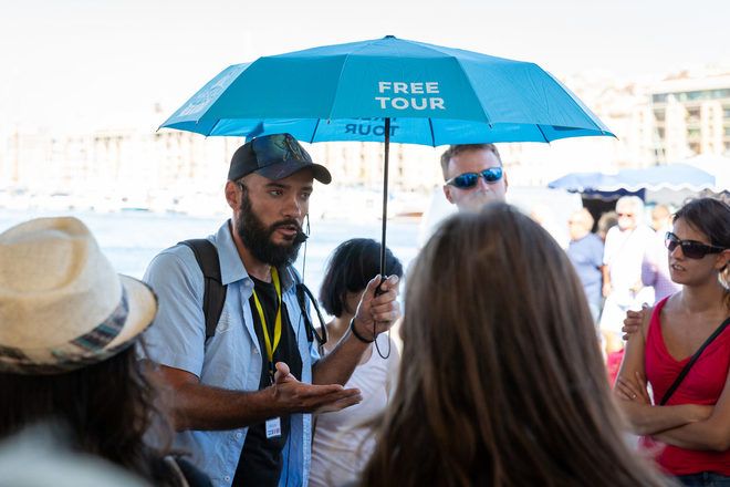 Guía de free tour con paragua en la mano explica algo a los viajeros via un microfono en el puerto de Marsella, Francia.